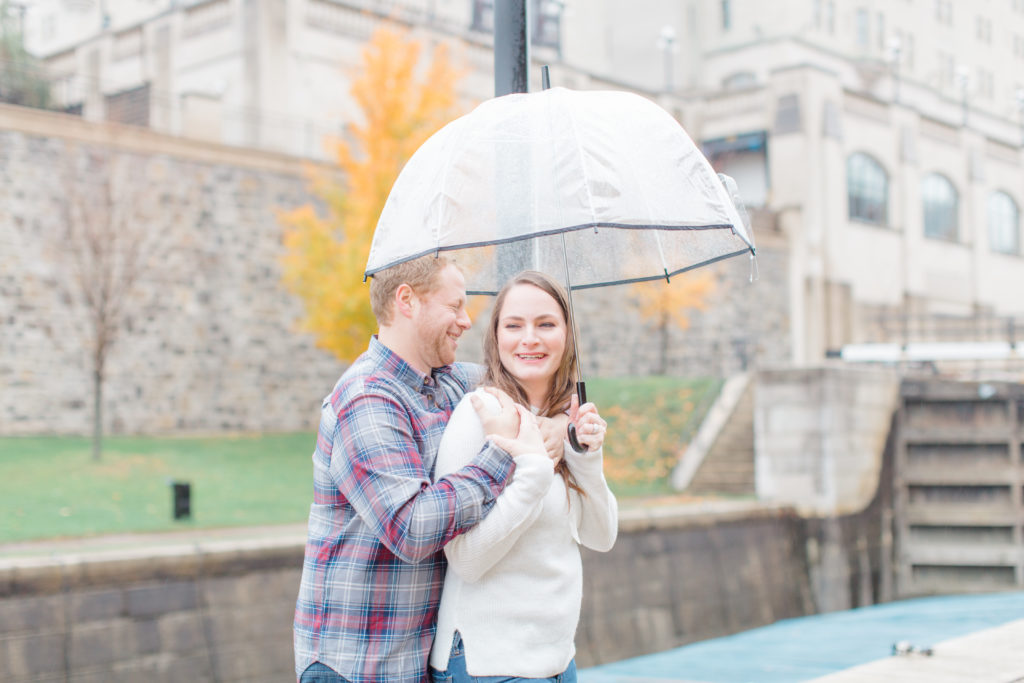Couple standing in the rain during engagement session with the Locks and Chateau Laurier in the background laughing 