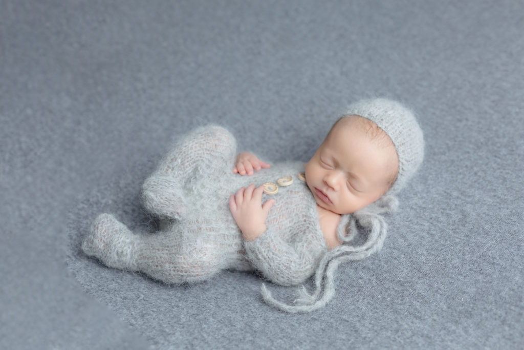 baby in a light grey knitted outfit with a hat and 3 buttons laying on back