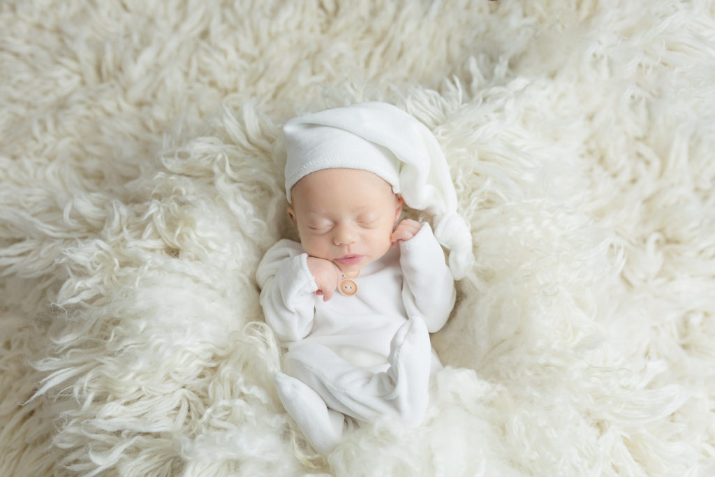baby lying in a white fluff with a white hat on a wool white rug 