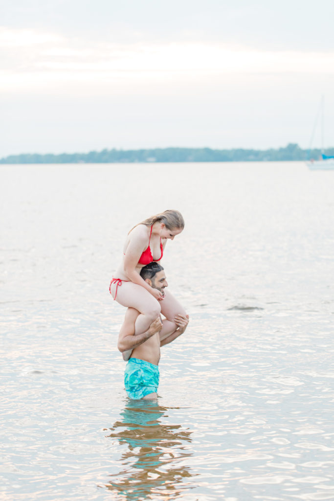 Cute Poses on the beach in Water - Ideas for what to wear for Engagement Photography, Modern Engagement Session Inspiration Wardrobe Ideas. Unsure of what to wear for your engagement photos, we've got you! Teal Board Shorts and Red Bikini -  Engagement at Britannia Beach, Nepean. Grey Loft Studio is Ottawa's Wedding and Engagement Photographer Videographer for Real couples, showcasing photos that are modern, bright, and fun.