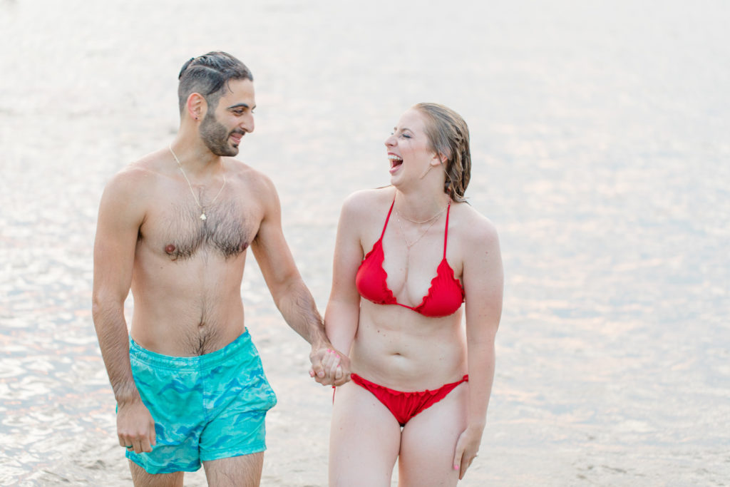 Cute Poses on the beach in Water - Ideas for what to wear for Engagement Photography, Modern Engagement Session Inspiration Wardrobe Ideas. Unsure of what to wear for your engagement photos, we've got you! Teal Board Shorts and Red Bikini -  Engagement at Britannia Beach, Nepean. Grey Loft Studio is Ottawa's Wedding and Engagement Photographer Videographer for Real couples, showcasing photos that are modern, bright, and fun.