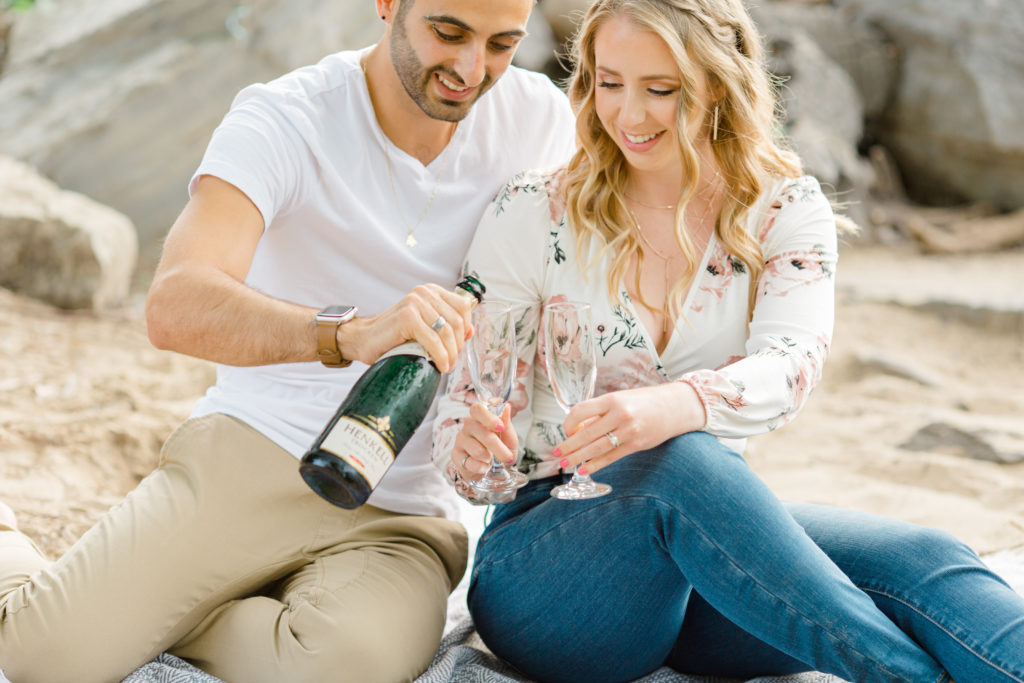 Pouring Champagne - Sitting on the Beach - Ideas for what to wear for Engagement Photography, Modern Engagement Session Inspiration Wardrobe Ideas. Unsure of what to wear for your engagement photos, we've got you! Romantic floral shirt & jeans.  White T-shirt & neutral pants . Boat Shoes and Fancy. beaded sandals. Engagement at Petrie Island, Orleans. Grey Loft Studio is Ottawa's Wedding and Engagement Photographer Videographer for Real couples, showcasing photos that are modern, bright, and fun.