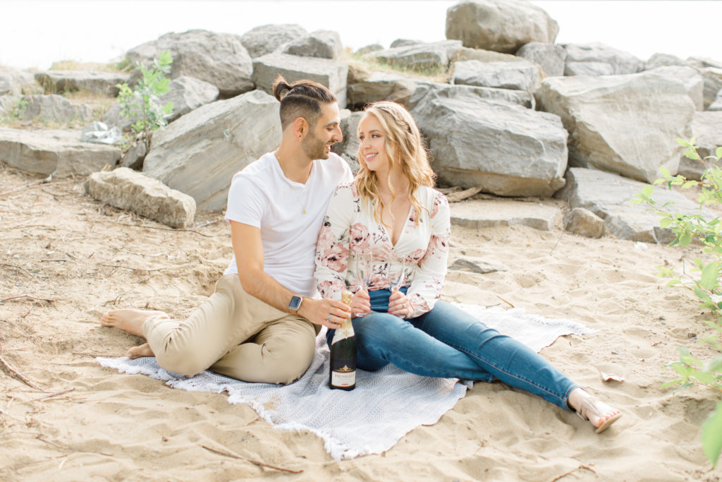 Drink the champagne - Sitting on the Beach - Ideas for what to wear for Engagement Photography, Modern Engagement Session Inspiration Wardrobe Ideas. Unsure of what to wear for your engagement photos, we've got you! Romantic floral shirt & jeans.  White T-shirt & neutral pants . Boat Shoes and Fancy. beaded sandals. Engagement at Petrie Island, Orleans. Grey Loft Studio is Ottawa's Wedding and Engagement Photographer Videographer for Real couples, showcasing photos that are modern, bright, and fun.