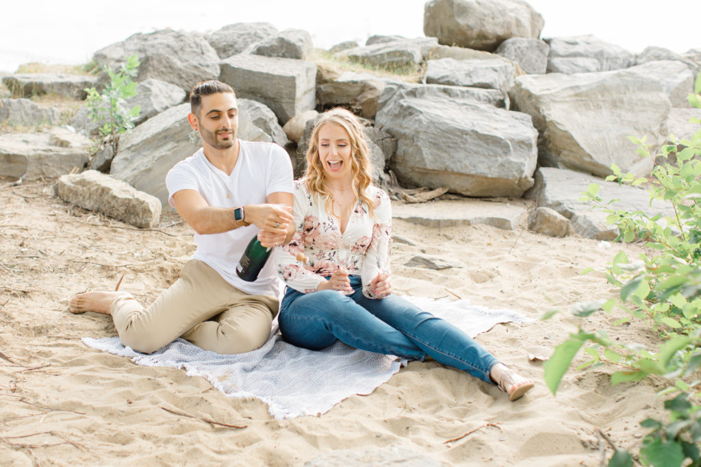 Popping the Champagne -Sitting on the Beach - Ideas for what to wear for Engagement Photography, Modern Engagement Session Inspiration Wardrobe Ideas. Unsure of what to wear for your engagement photos, we've got you! Romantic floral shirt & jeans.  White T-shirt & neutral pants . Boat Shoes and Fancy. beaded sandals. Engagement at Petrie Island, Orleans. Grey Loft Studio is Ottawa's Wedding and Engagement Photographer Videographer for Real couples, showcasing photos that are modern, bright, and fun.