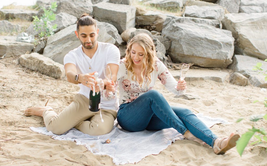 Champagne Spraying - Sitting on the Beach - Ideas for what to wear for Engagement Photography, Modern Engagement Session Inspiration Wardrobe Ideas. Unsure of what to wear for your engagement photos, we've got you! Romantic floral shirt & jeans.  White T-shirt & neutral pants . Boat Shoes and Fancy. beaded sandals. Engagement at Petrie Island, Orleans. Grey Loft Studio is Ottawa's Wedding and Engagement Photographer Videographer for Real couples, showcasing photos that are modern, bright, and fun.