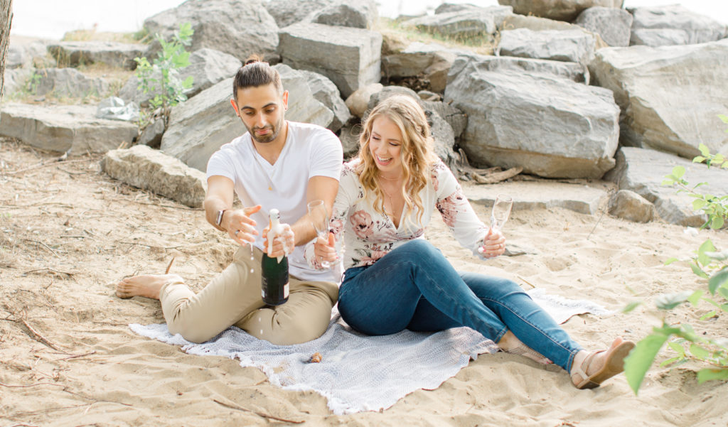 Champagne Spraying -  Sitting on the Beach - Ideas for what to wear for Engagement Photography, Modern Engagement Session Inspiration Wardrobe Ideas. Unsure of what to wear for your engagement photos, we've got you! Romantic floral shirt & jeans.  White T-shirt & neutral pants . Boat Shoes and Fancy. beaded sandals. Engagement at Petrie Island, Orleans. Grey Loft Studio is Ottawa's Wedding and Engagement Photographer Videographer for Real couples, showcasing photos that are modern, bright, and fun.