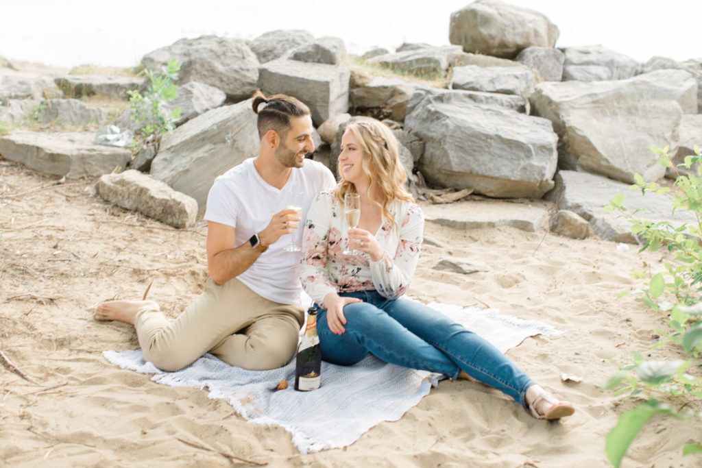 Cheers Shot - Sitting on the Beach - Ideas for what to wear for Engagement Photography, Modern Engagement Session Inspiration Wardrobe Ideas. Unsure of what to wear for your engagement photos, we've got you! Romantic floral shirt & jeans.  White T-shirt & neutral pants . Boat Shoes and Fancy. beaded sandals. Engagement at Petrie Island, Orleans. Grey Loft Studio is Ottawa's Wedding and Engagement Photographer Videographer for Real couples, showcasing photos that are modern, bright, and fun.