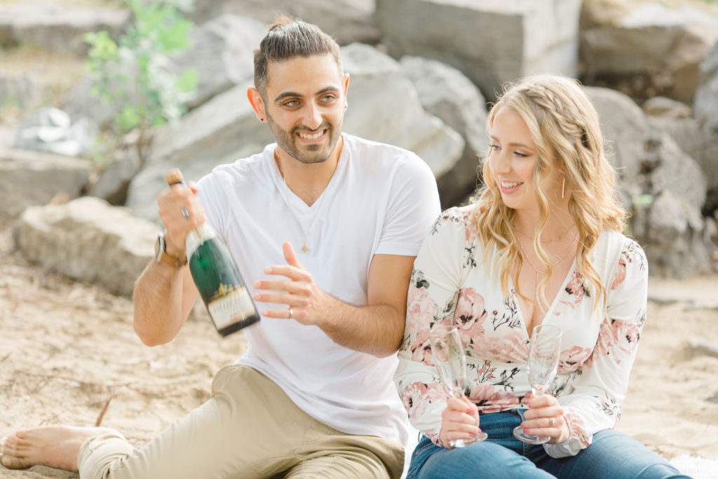Shake up the Champagne - Sitting on the Beach - Ideas for what to wear for Engagement Photography, Modern Engagement Session Inspiration Wardrobe Ideas. Unsure of what to wear for your engagement photos, we've got you! Romantic floral shirt & jeans.  White T-shirt & neutral pants . Boat Shoes and Fancy. beaded sandals. Engagement at Petrie Island, Orleans. Grey Loft Studio is Ottawa's Wedding and Engagement Photographer Videographer for Real couples, showcasing photos that are modern, bright, and fun.