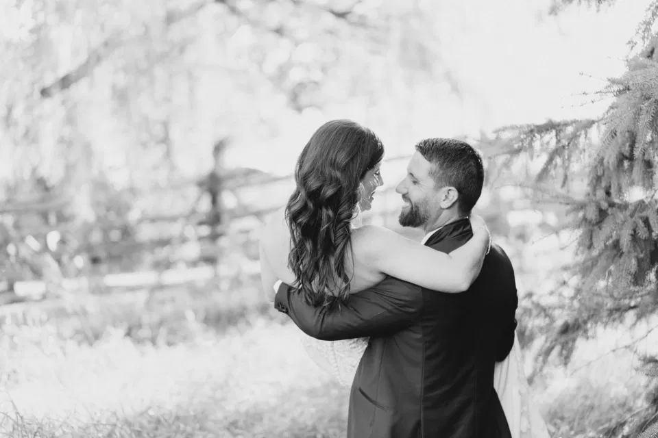 Lifting his Bride - Black and White -  Bride and Groom - Standing in a Field - Off-White, Silver, Greenery. Eucalyptus Leaves. An inspiration filled with soft neutrals, lush florals, and layers of romantic textures all set at Evermore Weddings and Events, Almonte Ontario. 
