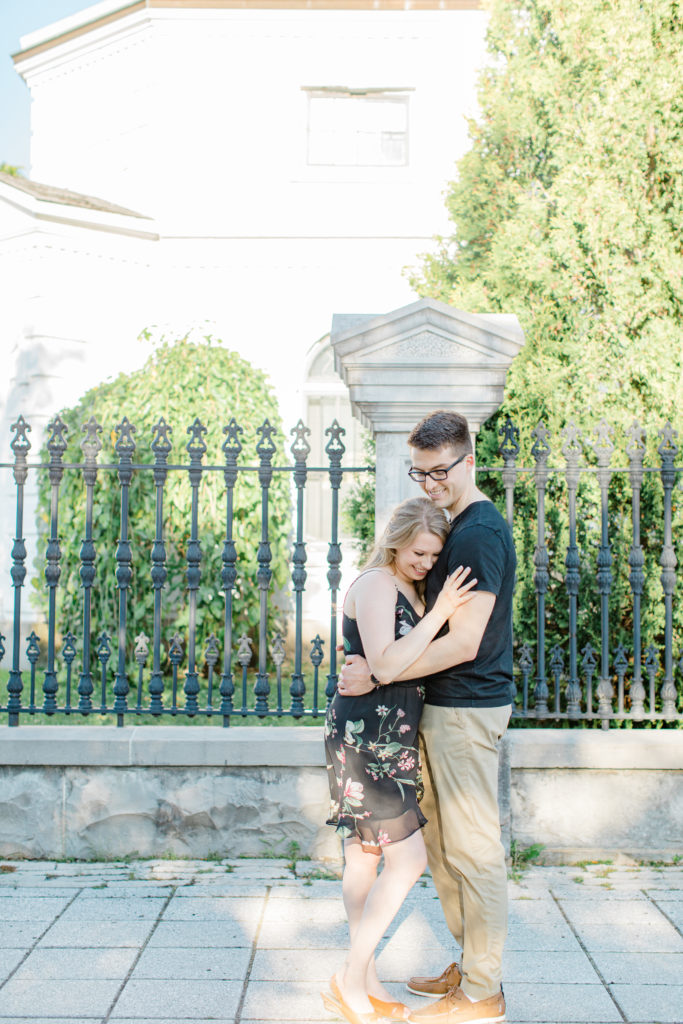 Cuddles while Standing at Rideau Hall - Ideas for what to wear for Engagement Photography, Modern Engagement Session Inspiration Wardrobe Ideas. Unsure of what to wear for your engagement photos, we've got you! Romantic floral dress. Black Polo T-shirt & neutral pants . Boat Shoes and Fancy beaded wedges. Engagement downtown Ottawa. Grey Loft Studio is Ottawa's Wedding and Engagement Photographer Videographer for Real couples, showcasing photos that are modern, bright, and fun.