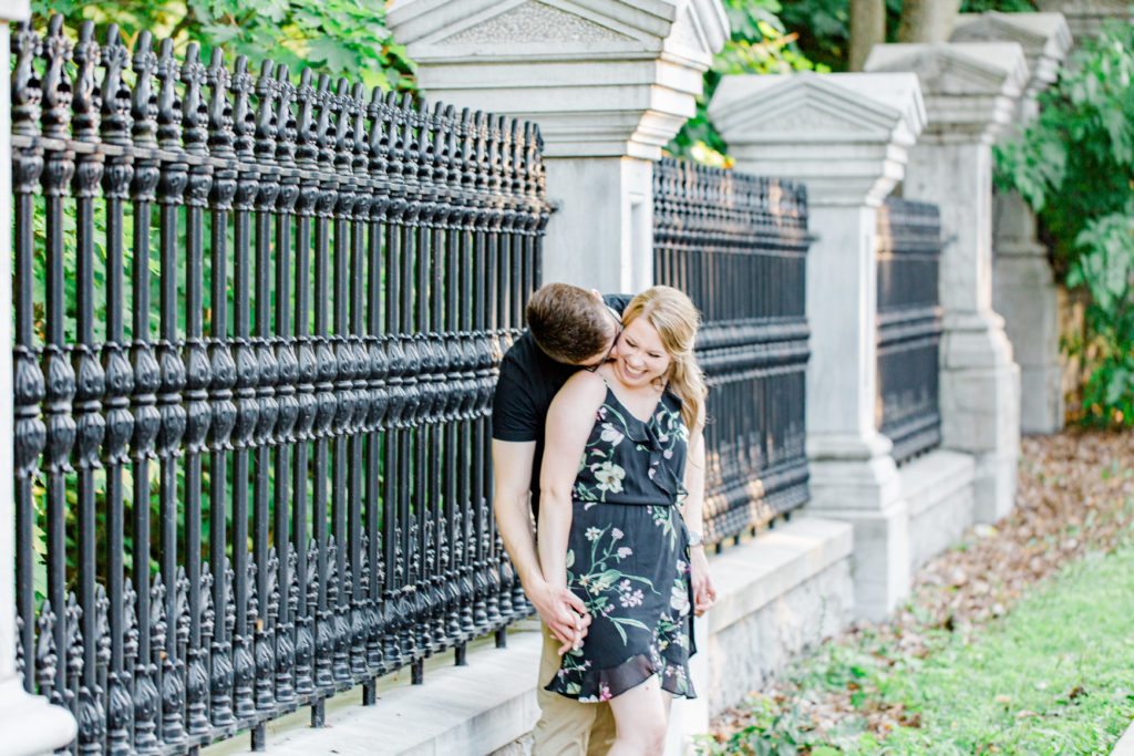 Kissing the Neck - Holding Hands Shot - Engagement Photo Poses- Posing while at Rideau Hall - Ideas for what to wear for Engagement Photography, Modern Engagement Session Inspiration Wardrobe Ideas. Unsure of what to wear for your engagement photos, we've got you! Romantic floral dress. Black Polo T-shirt & neutral pants . Boat Shoes and Fancy beaded wedges. Engagement downtown Ottawa. Grey Loft Studio is Ottawa's Wedding and Engagement Photographer Videographer for Real couples, showcasing photos that are modern, bright, and fun.