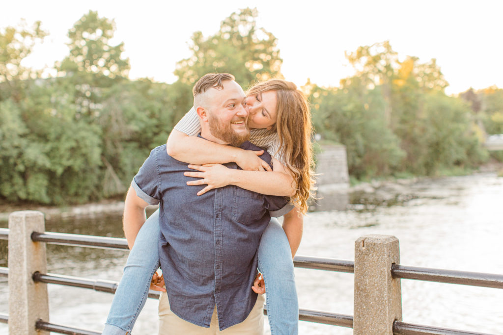 Kissing during piggy back pose - Watson's Mill Engagement Session Manotick - Bright & Airy photography - Grey Loft Studio - Ottawa Wedding Photographer - Ottawa Wedding Videographer - Engagement Session Locations in Ottawa - Summer Engagement session - Light blue and Cream with casual jeans and strap sandals. Ottawa Photo Studio.