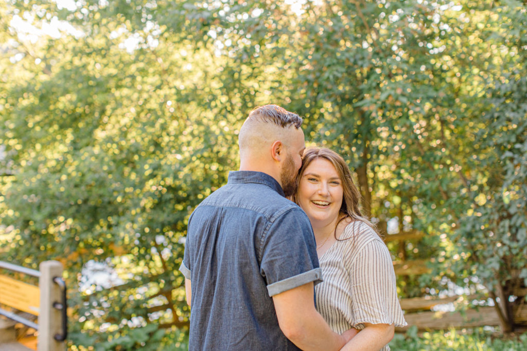 Couple holding each other close and walking looking back -engagement session - Watson's Mill Engagement Session Manotick - Bright & Airy photography - Grey Loft Studio - Ottawa Wedding Photographer - Ottawa Wedding Videographer - Engagement Session Locations in Ottawa - Summer Engagement session - Light blue and Cream with casual jeans and strap sandals. Ottawa Photo Studio.