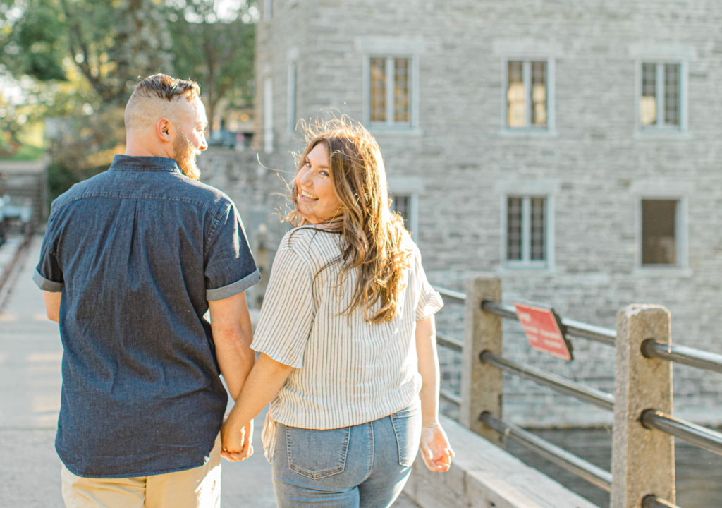Couple holding each other close and walking looking back -engagement session - Watson's Mill Engagement Session Manotick - Bright & Airy photography - Grey Loft Studio - Ottawa Wedding Photographer - Ottawa Wedding Videographer - Engagement Session Locations in Ottawa - Summer Engagement session - Light blue and Cream with casual jeans and strap sandals. Ottawa Photo Studio.