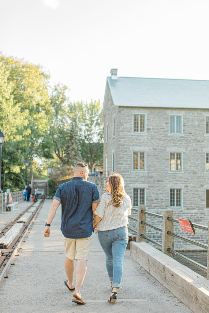 Couple holding each other close and walking -engagement session - Watson's Mill Engagement Session Manotick - Bright & Airy photography - Grey Loft Studio - Ottawa Wedding Photographer - Ottawa Wedding Videographer - Engagement Session Locations in Ottawa - Summer Engagement session - Light blue and Cream with casual jeans and strap sandals. Ottawa Photo Studio.