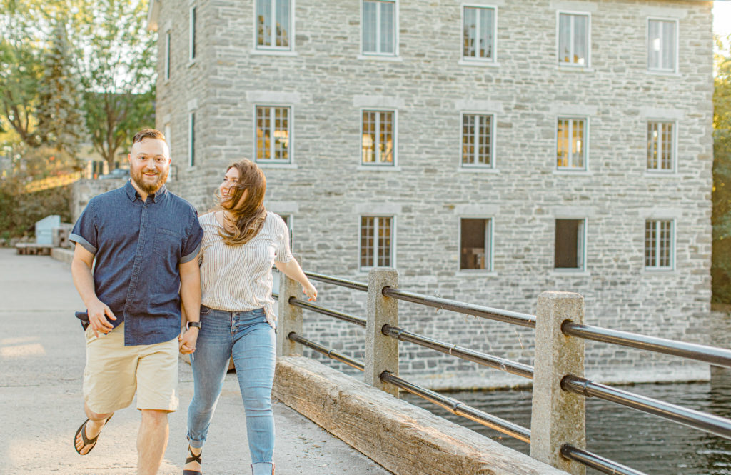 Couple holding each other close and walking-engagement session - Watson's Mill Engagement Session Manotick - Bright & Airy photography - Grey Loft Studio - Ottawa Wedding Photographer - Ottawa Wedding Videographer - Engagement Session Locations in Ottawa - Summer Engagement session - Light blue and Cream with casual jeans and strap sandals. Ottawa Photo Studio.
