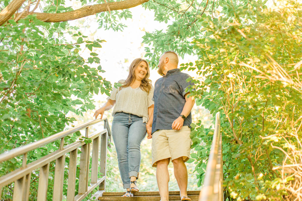 Walking down the stairs at an engagement session - Couple holding each other close -engagement session - Watson's Mill Engagement Session Manotick - Bright & Airy photography - Grey Loft Studio - Ottawa Wedding Photographer - Ottawa Wedding Videographer - Engagement Session Locations in Ottawa - Summer Engagement session - Light blue and Cream with casual jeans and strap sandals. Ottawa Photo Studio.