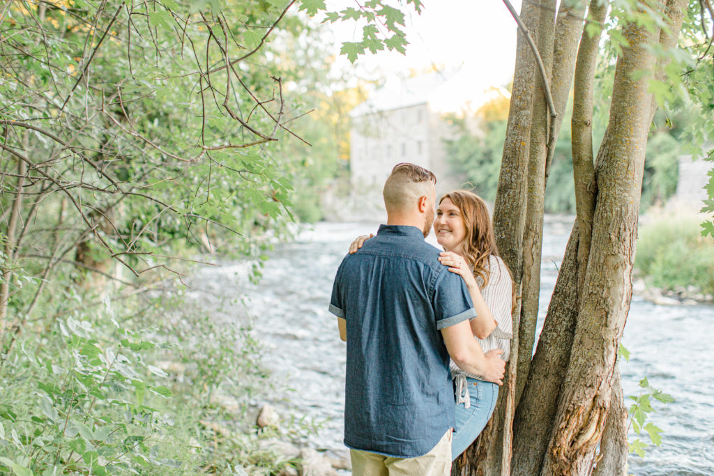 Look at each other and smile - by the water - Watson's Mill Engagement Session Manotick - Bright & Airy photography - Grey Loft Studio - Ottawa Wedding Photographer - Ottawa Wedding Videographer - Engagement Session Locations in Ottawa - Summer Engagement session - Light blue and Cream with casual jeans and strap sandals. Ottawa Photo Studio.