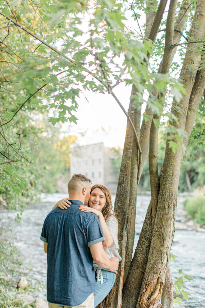 Look at the camera over the shoulder - hold the waist - Watson's Mill Engagement Session Manotick - Bright & Airy photography - Grey Loft Studio - Ottawa Wedding Photographer - Ottawa Wedding Videographer - Engagement Session Locations in Ottawa - Summer Engagement session - Light blue and Cream with casual jeans and strap sandals. Ottawa Photo Studio.