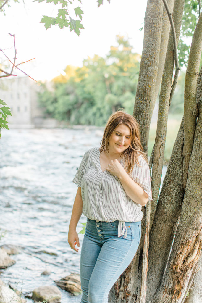woman leaning on tree near water - Watson's Mill Engagement Session Manotick - Bright & Airy photography - Grey Loft Studio - Ottawa Wedding Photographer - Ottawa Wedding Videographer - Engagement Session Locations in Ottawa - Summer Engagement session - Light blue and Cream with casual jeans and strap sandals. Ottawa Photo Studio.