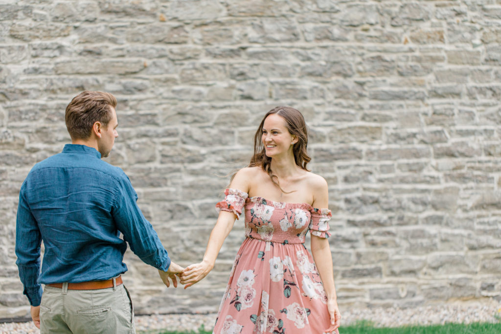 couple spinning near bricks -Ideas for what to wear for Engagement Photography, Modern Engagement Session Inspiration Wardrobe Ideas. Unsure of what to wear for your engagement photos, we've got you! Romantic white with Pink Flower dress for Summer Engagement in Ottawa. Grey Loft Studio is Ottawa's Wedding and Engagement Photographer for Real couples, showcasing photos that are modern, bright, and fun. Pinhey's Point Dunrobin. Light and Airy Photography Videography Team