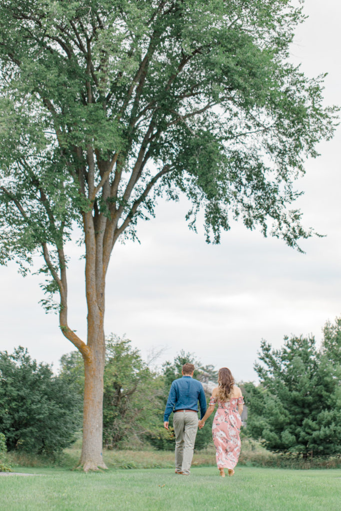 Couple walking near large tree -Ideas for what to wear for Engagement Photography, Modern Engagement Session Inspiration Wardrobe Ideas. Unsure of what to wear for your engagement photos, we've got you! Romantic white with Pink Flower dress for Summer Engagement in Ottawa. Grey Loft Studio is Ottawa's Wedding and Engagement Photographer for Real couples, showcasing photos that are modern, bright, and fun. Pinhey's Point Dunrobin.